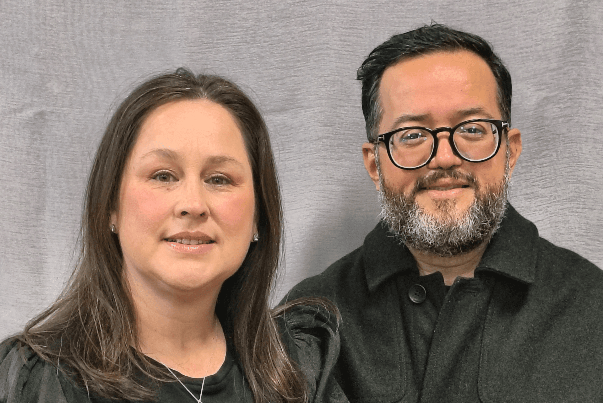 Two people smiling, one wearing glasses, in front of a gray backdrop.