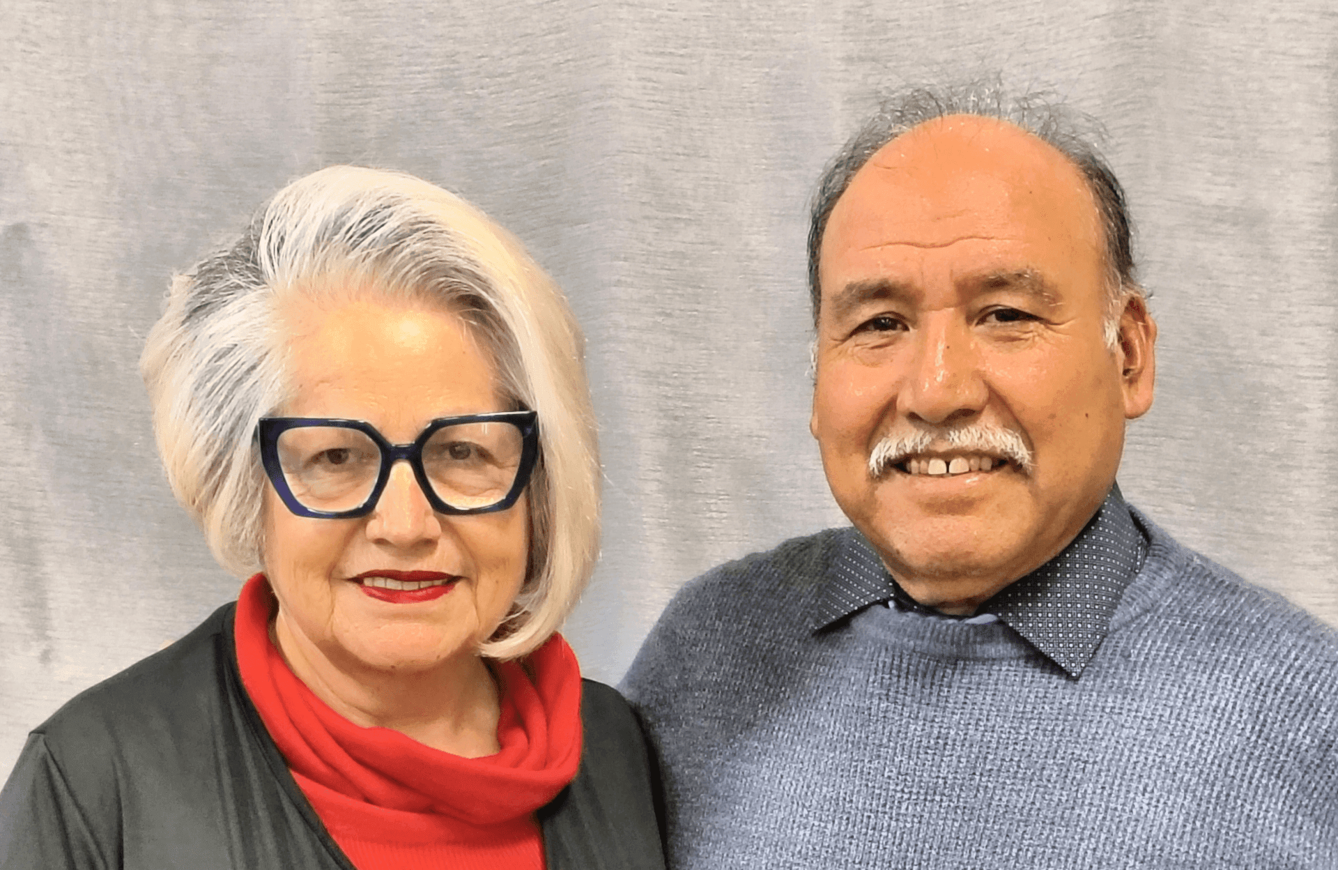 A smiling older couple standing together in front of a neutral gray background.