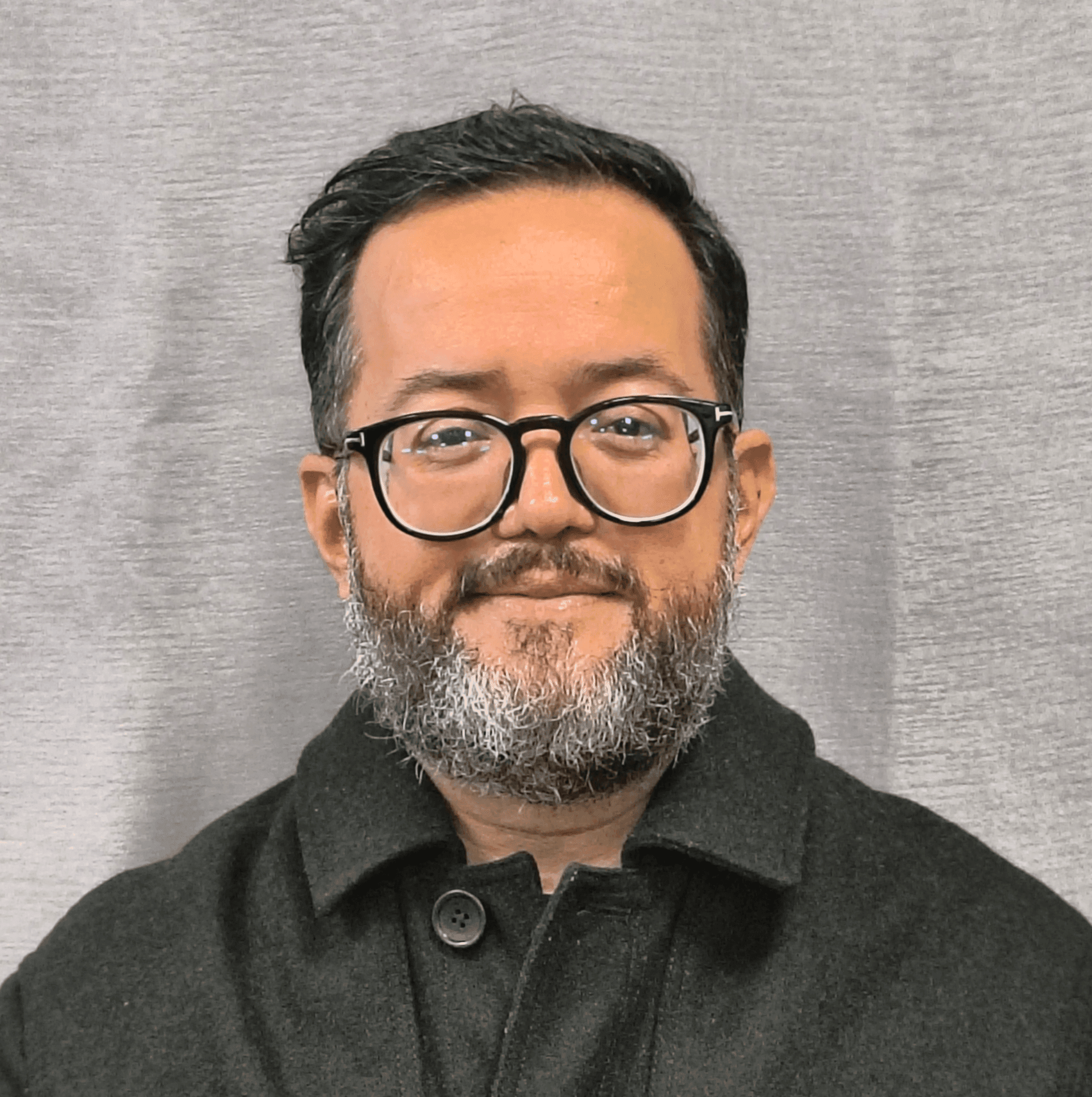 Man with glasses and a beard wearing a dark coat against a gray textured background.