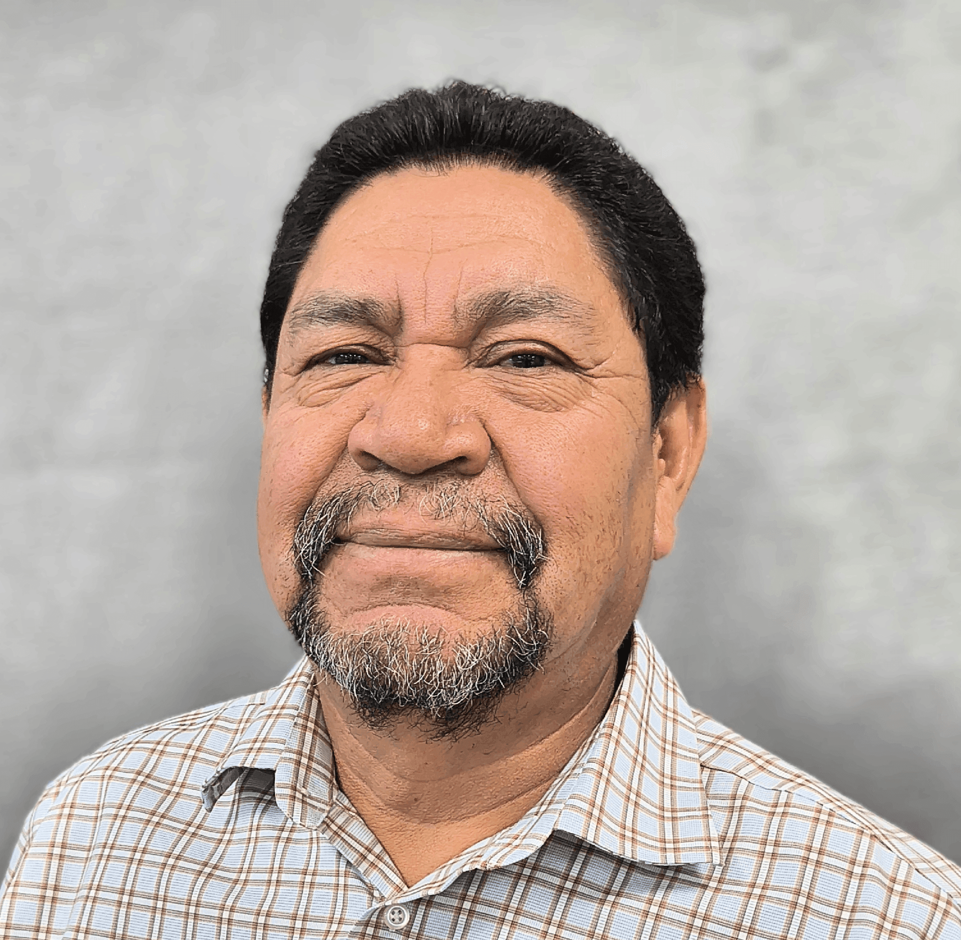 Man with short dark hair and a beard smiling, wearing a plaid shirt against a neutral background.